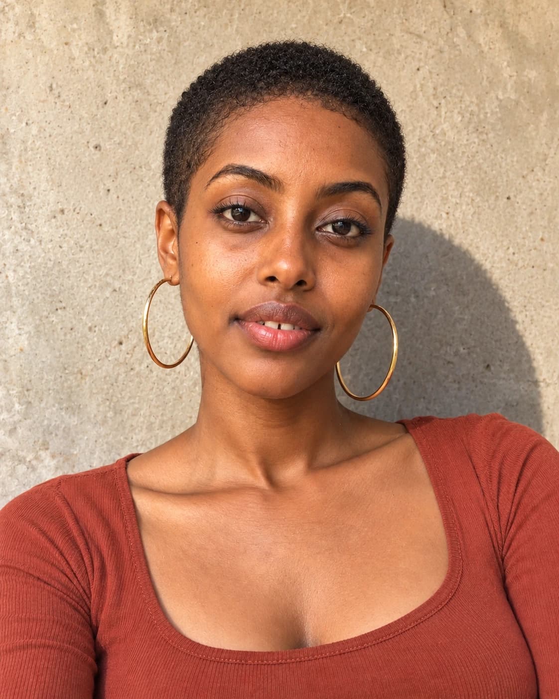 Woman in her late 20s with a #2 buzz cut on coily hair and a soft rounded hairline, close portrait with large gold hoops against a sunlit concrete wall