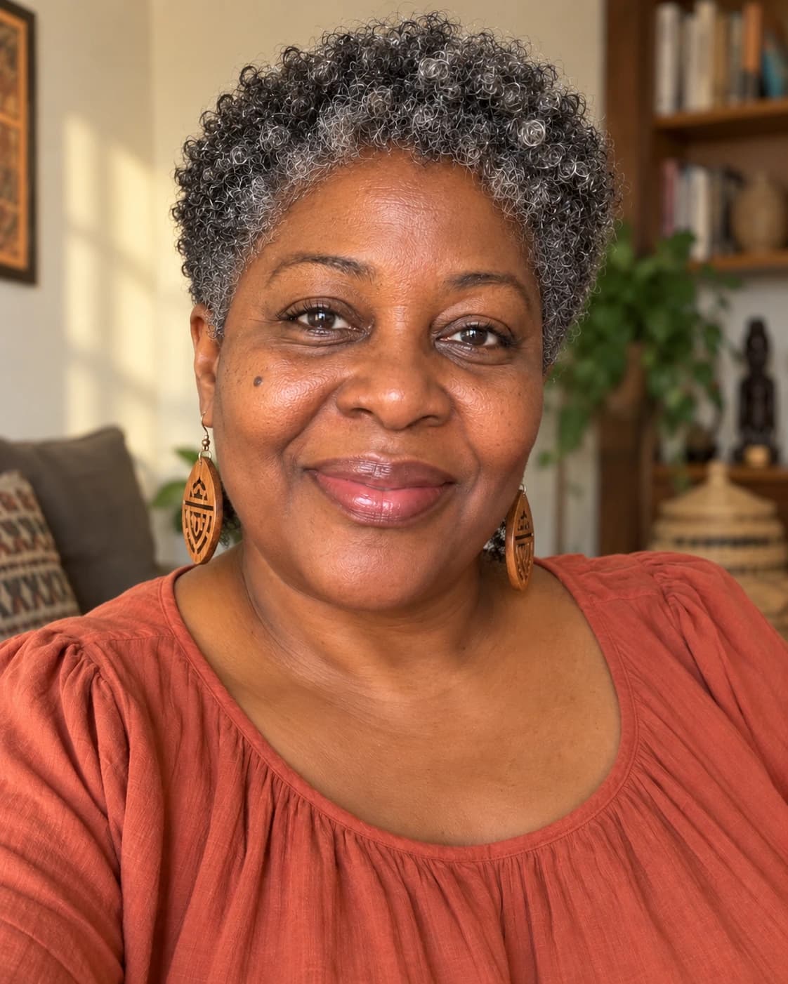 Woman in her late 50s with a tapered coily pixie, defined coils on top with natural gray and short tapered sides, three-quarter view in a warm living room
