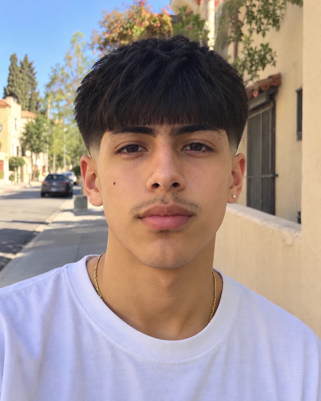 Young man in his 20s with an Edgar cut, a straight blunt fringe across the forehead over a high taper, front view on a sunny street