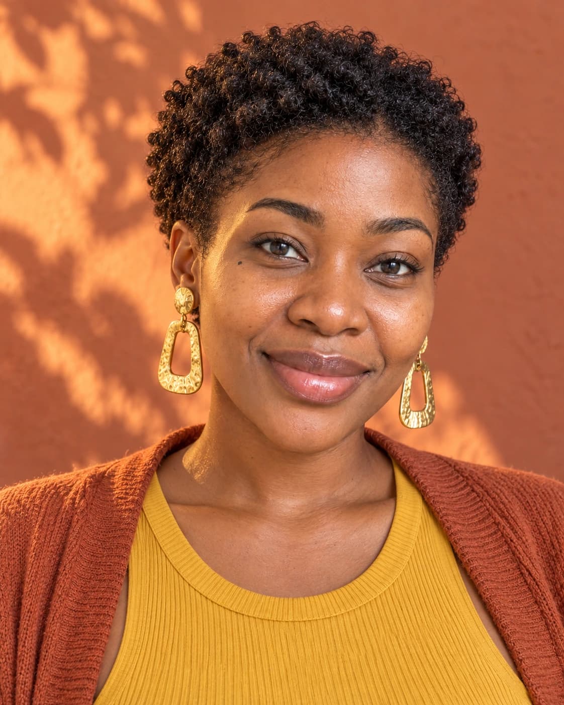 Woman in her 30s with a tapered teeny weeny afro, defined coils on top and short sides, front view against a terracotta wall in golden light
